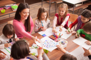 Students boys and girls sitting together around the table in clasroom and drawing. With them is their young and beautiful teacher. She teaches children and is smiling
